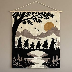 Può includere: Un arazzo rettangolare all'uncinetto che presenta la sagoma nera di un gruppo di nove figure che camminano attraverso un paesaggio. La scena include un grande albero, montagne, un sole dorato e un riflesso nell'acqua, appeso a un'asta di legno.