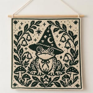 May include: A square cross-stitch wall hanging featuring a dark green frog wearing a wizard hat with a crescent moon and stars. The frog is framed by a border of dark green leaves, bell-shaped flowers, and mushrooms on a cream-colored fabric.