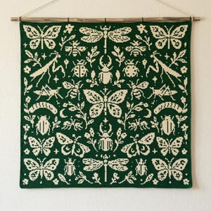 May include: A square tapestry featuring a dark green background with a cream-colored grid pattern of various insects including butterflies, dragonflies, beetles, praying mantises, and bees, all suspended from a wooden rod with a string.