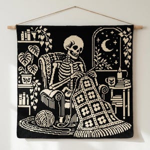 May include: A black and cream crochet wall hanging featuring a skeleton sitting in a chair, knitting a floral blanket. The scene includes a potted plant, a bookshelf, a side table with a mug and candle, and a window showing a crescent moon.