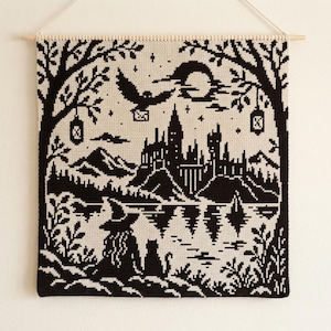 May include: A black and cream wall hanging featuring a cross-stitch style design of a castle on a hill by a lake. The scene includes a figure in a pointed hat, a cat, a flying owl carrying a letter, trees, lanterns, and a crescent moon.