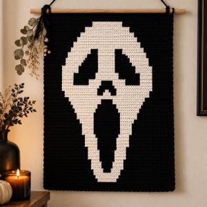 Könnte beinhalten: Ein rechteckiger Wandbehang aus Häkelarbeit mit einem pixeligen, cremeweißen Geistermasken-Motiv auf schwarzem Hintergrund. Das Textil ist an einem hellen Holzstab mit einer schwarzen Kordel befestigt und erzeugt einen starken Kontrast.