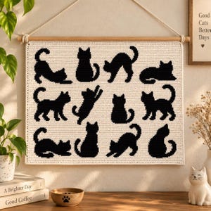 May include: A rectangular cream-colored crochet wall hanging featuring twelve black cat silhouettes in various poses. The textile is suspended from a wooden dowel with a white rope. The cats are arranged in a grid pattern across the fabric surface.