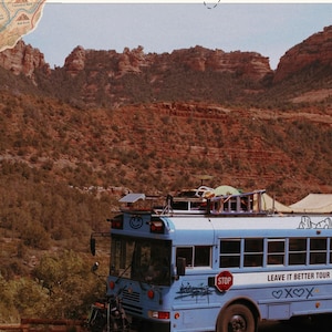 May include: A light blue bus parked in front of red rock mountains. The bus features a roof rack, a front-mounted bicycle, and text that reads Arizona Travel Guide, Leave It Better Tour, and various symbols including a smiley face and hearts.