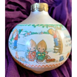 Könnte beinhalten: Eine runde, weiße Weihnachtskugel aus Glas mit einer farbenfrohen Illustration von Kindern in einer winterlichen Szene. Das Design zeigt Figuren beim Schlittschuhlaufen, auf einer Leiter und am Lagerfeuer. Der Text lautet Happy is the joy of bringing home the Christmas tree.
