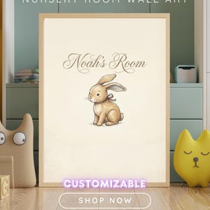May include: A rectangular wall art print in a light wood frame featuring a beige background with a brown illustration of a seated rabbit wearing a small ribbon. The text "Noah's Room" is printed in a brown script font above the rabbit illustration.