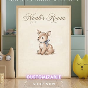 Op de afbeelding: Een rechthoekige wandkunstprint in een lichthouten lijst met een beige achtergrond en de tekst Noah's Room in een donkerbruin scriptlettertype. Onder de tekst staat een aquarelillustratie van een klein bruin reekalfje met een blauw lint.