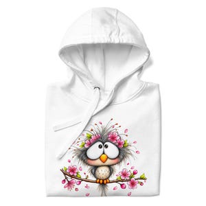 Peut inclure: Un sweat à capuche blanc orné d'une illustration d'un petit oiseau aux grands yeux, au plumage gris ébouriffé et portant des fleurs roses sur la tête, perché sur une branche brune entourée de fleurs de cerisier roses et de pétales.