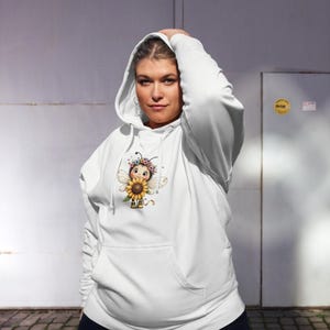 Peut inclure: Un sweat-shirt à capuche blanc arborant un motif central représentant une abeille de dessin animé portant une couronne de fleurs colorées et tenant un grand tournesol jaune. Le sweat possède une poche kangourou et des manches longues.