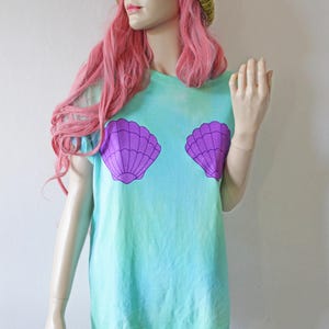 Könnte beinhalten: Ein türkisfarbenes Batik-T-Shirt mit einem lila Muschel-Motiv. Das Shirt hat eine lockere Passform und wird mit einer senfgelben Strickmütze getragen.