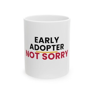 May include: A white ceramic mug featuring the black text EARLY ADOPTER on the top line, a thin horizontal yellow line in the middle, and the red text NOT SORRY on the bottom line. The mug has a smooth, cylindrical body and a clean white finish.