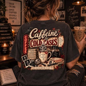 May include: A charcoal gray t-shirt featuring a graphic print on the back with the text Caffeine & Cold Cases. The design includes a coffee mug, a stack of books, a magnifying glass, a raven, and a coffee pot, all surrounded by red accents.