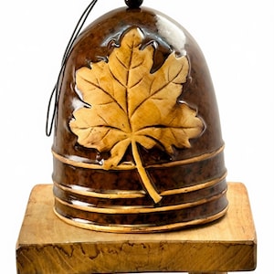 May include: A bell-shaped ceramic ornament with a glossy dark brown mottled glaze. The front features a large, raised, light tan maple leaf relief. Three horizontal ridges circle the base, and a black cord is attached to the top bead.
