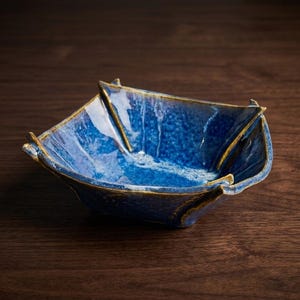 May include: A square ceramic bowl with a deep blue, mottled glaze and a glossy finish. The bowl features four distinct, raised, pointed corners and a dark, textured rim that contrasts with the vibrant blue interior and exterior surfaces.