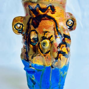 May include: A ceramic vase with a textured, uneven surface featuring a glossy finish. The upper portion is a mottled blend of gold, brown, and black, while the lower section is a vibrant, solid blue. It has two side handles with holes.
