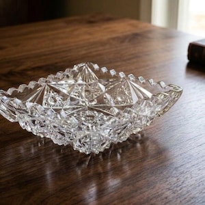 May include: A square clear glass dish featuring a faceted geometric pattern with sharp edges and a star-like design in the center. The glass has a textured, pressed surface with multiple triangular and diamond-shaped facets throughout.