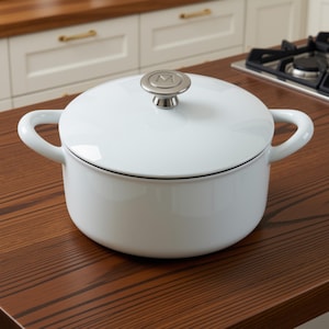 May include: A round white enameled Dutch oven with two side handles and a matching lid. The lid features a silver-toned metallic knob with a circular letter M embossed on the top. The pot has a smooth, glossy finish and a clean, minimalist design.