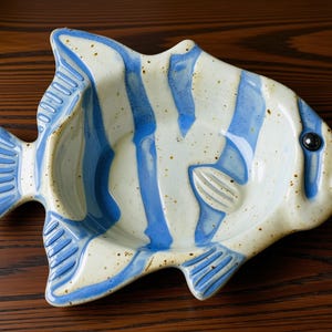 May include: A ceramic fish-shaped dish featuring a cream-colored base with speckled brown dots and vertical blue stripes. The dish has a molded tail, fins, and a single dark eye detail, creating a functional bowl with a distinct aquatic silhouette.