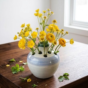 May include: A round, ceramic flower frog vase with a white and light blue glazed finish, featuring multiple circular openings at the top. The vase is filled with a vibrant arrangement of bright yellow ranunculus and small white daisy-like flowers.