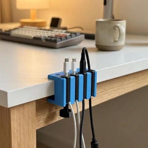 May include: A blue and black plastic cable organiser clamp attached to the edge of a white desk. The device features four vertical slots holding two white charging cables and one black braided cable, secured by a black tightening knob underneath.