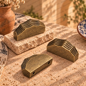 May include: Three antique brass-toned metal drawer pulls with a stepped, mountain-like silhouette. Each handle features a textured, layered design on the top edge and two circular screw holes on the flat front face for mounting to furniture.