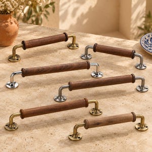 May include: A collection of six cabinet pulls featuring cylindrical dark brown wood handles mounted on metal brackets. The hardware includes both brushed brass and polished silver finishes, each with circular base plates and visible screw holes.