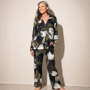May include: A two-piece pajama set featuring a long-sleeved button-down shirt and matching trousers. The fabric is black with a repeating botanical print of large white magnolia flowers and dark green leaves throughout the entire garment.