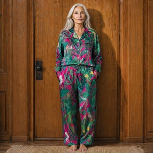 May include: A long-sleeved button-down pajama shirt and matching loose-fitting trousers featuring a vibrant tropical print with green foliage, yellow accents, and white bird motifs set against a deep magenta background with black piping details.