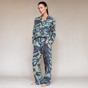 May include: A two-piece pajama set featuring a long-sleeved button-down shirt with a collar and matching straight-leg trousers. The set is light green and decorated with a repeating dark blue and yellow bird-like pattern throughout the fabric.