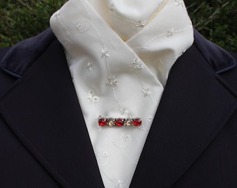 May include: A white, embroidered stock tie with a red jeweled pin. The stock tie has a delicate floral pattern and is worn with a dark blue jacket.