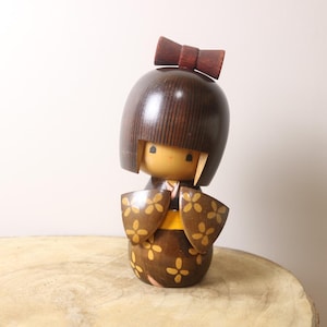 以下が含まれることがあります： 赤いリボンをつけたダークブラウンのボブヘアの木製こけし人形。顔は明るい色で、シンプルな黒い目と小さな口が描かれています。黄色い花の模様が繰り返されたダークブラウンの着物を着ています。