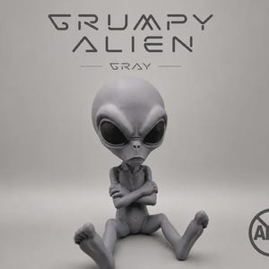 Puede incluir: Una figura de alienígena gris con cabeza grande, ojos ovalados oscuros y boca fruncida, sentada con los brazos cruzados. El texto GRUMPY ALIEN GRAY aparece arriba y un logotipo circular de AMS es visible en la esquina inferior derecha.
