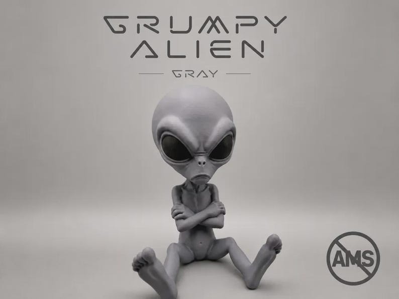 Puede incluir: Una figura de alien&iacute;gena gris con cabeza grande, ojos ovalados oscuros y boca fruncida, sentada con los brazos cruzados. El texto GRUMPY ALIEN GRAY aparece arriba y un logotipo circular de AMS es visible en la esquina inferior derecha.