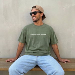 May include: An olive green short-sleeve t-shirt featuring the text ALGORITHMIC TRADER printed in white across the chest. The shirt is paired with light blue denim jeans, a tan baseball cap, and dark-framed sunglasses with black lenses.