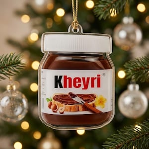 May include: A flat, clear acrylic Christmas ornament shaped like a jar of chocolate hazelnut spread. The label features the text Kheyri in bold black and red letters, an illustration of a slice of bread with spread, and a small yellow flower.