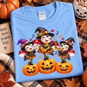 Könnte beinhalten: Ein hellblaues T-Shirt mit einer Grafik von drei Comic-Marienkäfern, die Hexenhüte und orangefarbene Röcke tragen und jeweils auf einem Kürbis stehen. Die Marienkäfer spielen Gitarre, Tamburin und Trompete, umgeben von Musiknoten.