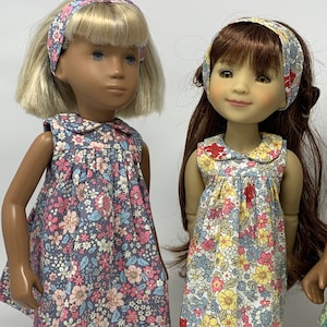 May include: Two dolls wearing floral print dresses and matching headbands. The dresses feature a variety of colorful flowers on a dark background. One doll has blonde hair, and the other has brown hair. The dolls are wearing white socks and black shoes.