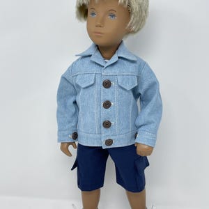 May include: A doll wearing a light blue denim jacket with brown buttons and dark blue cargo shorts. The doll has blonde hair and is wearing gray and white sneakers.