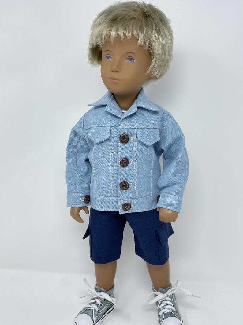 May include: A doll wearing a light blue denim jacket with brown buttons and dark blue cargo shorts. The doll has blonde hair and is wearing gray and white sneakers.