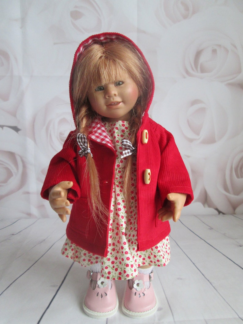 Pu&ograve; includere: Una bambola con lunghi capelli biondi indossa una giacca rossa in velluto a coste con cappuccio e alamari, un vestito bianco con un piccolo motivo floreale rosso e scarpe rosa chiaro con decorazioni a fiore bianco. Il cappuccio &egrave; foderato a quadretti.