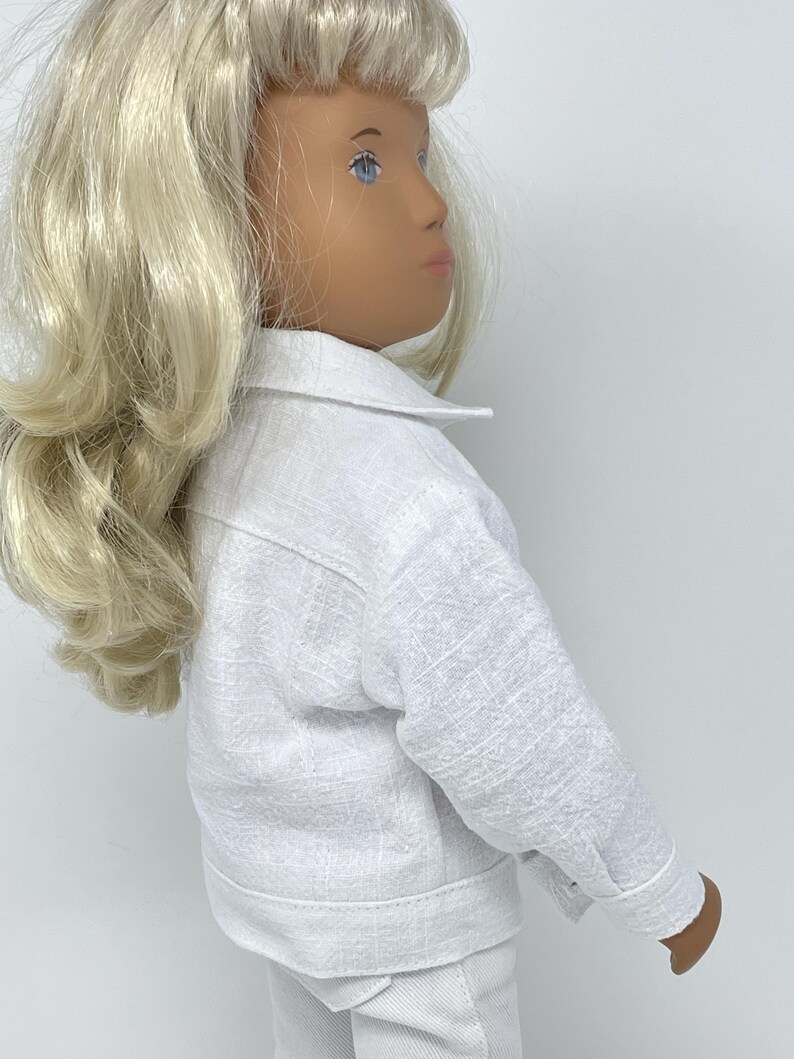 May include: A white doll wearing a white jacket and pants. The jacket has a collar and buttoned cuffs. The doll has blonde hair and blue eyes. The outfit appears to be made of a textured fabric.