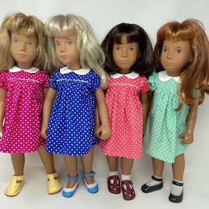 Puede incluir: Cuatro muñecas con diferentes colores de cabello: rubio, rubio claro, castaño oscuro y rojo. Cada muñeca viste un vestido de lunares en rosa, azul, coral y verde menta, con cuellos blancos. Llevan zapatos a juego en amarillo, azul, burdeos y negro.