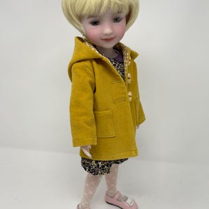 Pu&ograve; includere: Una bambola con capelli biondi corti indossa un cappotto con cappuccio in velluto a coste giallo senape con chiusure a levetta, un vestito scuro a fiori con colletto viola, collant bianchi a motivi e scarpe rosa chiaro con gattini.