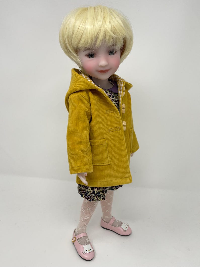 Pu&ograve; includere: Una bambola con capelli biondi corti indossa un cappotto con cappuccio in velluto a coste giallo senape con chiusure a levetta, un vestito scuro a fiori con colletto viola, collant bianchi a motivi e scarpe rosa chiaro con gattini.
