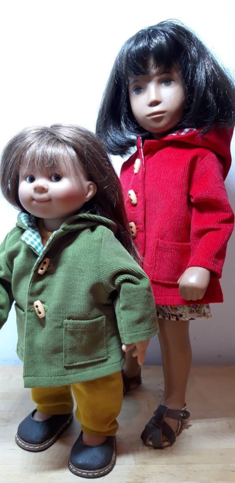 Pu&ograve; includere: Due bambole in piedi una accanto all'altra. La bambola pi&ugrave; bassa ha i capelli castani, una giacca verde in velluto a coste con cappuccio e bottoni in legno, pantaloni gialli e scarpe nere. La pi&ugrave; alta ha i capelli neri e una giacca rossa.