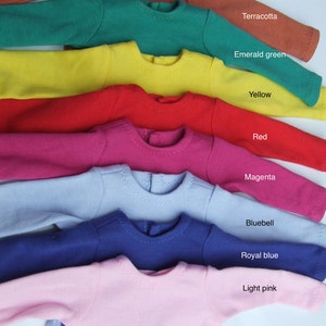 May include: A stack of eight long-sleeved shirts in different colors, including terracotta, emerald green, yellow, red, magenta, bluebell, royal blue, and light pink. The shirts are arranged from darkest to lightest.
