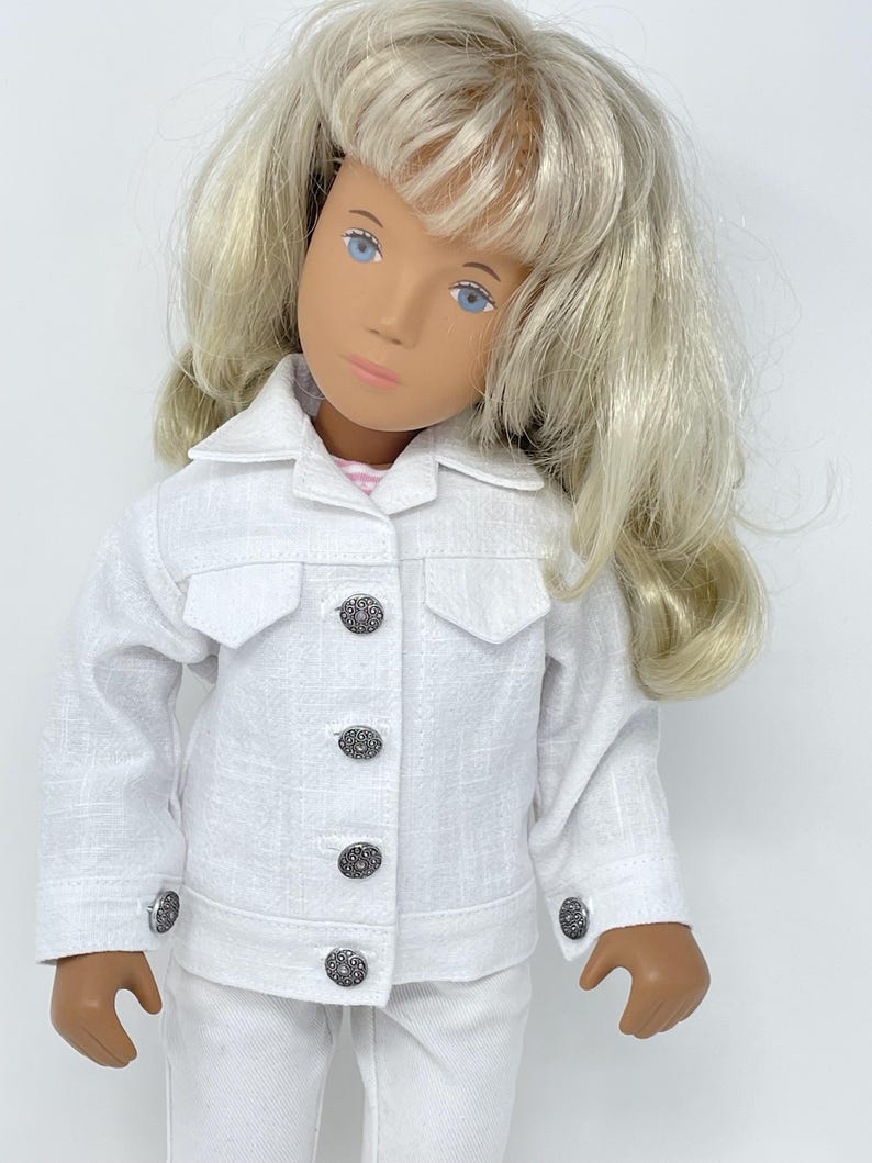 May include: A doll wearing a white jacket and matching pants. The jacket has decorative buttons and pocket flaps. The doll has blonde hair and blue eyes. The outfit is made of a textured fabric.