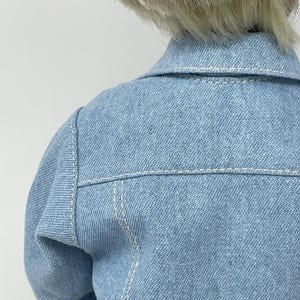 May include: A light blue denim jacket with white stitching details. The jacket features a classic collar and is designed with a tailored fit. The back of the jacket is visible, showcasing the shoulder and upper back area.
