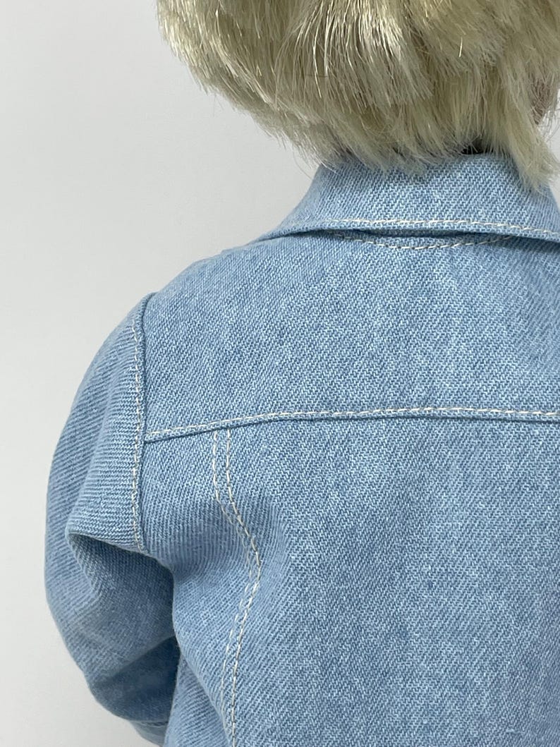 May include: A light blue denim jacket with white stitching details. The jacket features a classic collar and is designed with a tailored fit. The back of the jacket is visible, showcasing the shoulder and upper back area.