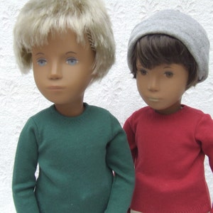 Può includere: Due bambole che indossano maglioni. Una bambola ha i capelli biondi e indossa un maglione verde. L'altra bambola ha i capelli castani e indossa un maglione rosso e un berretto grigio.